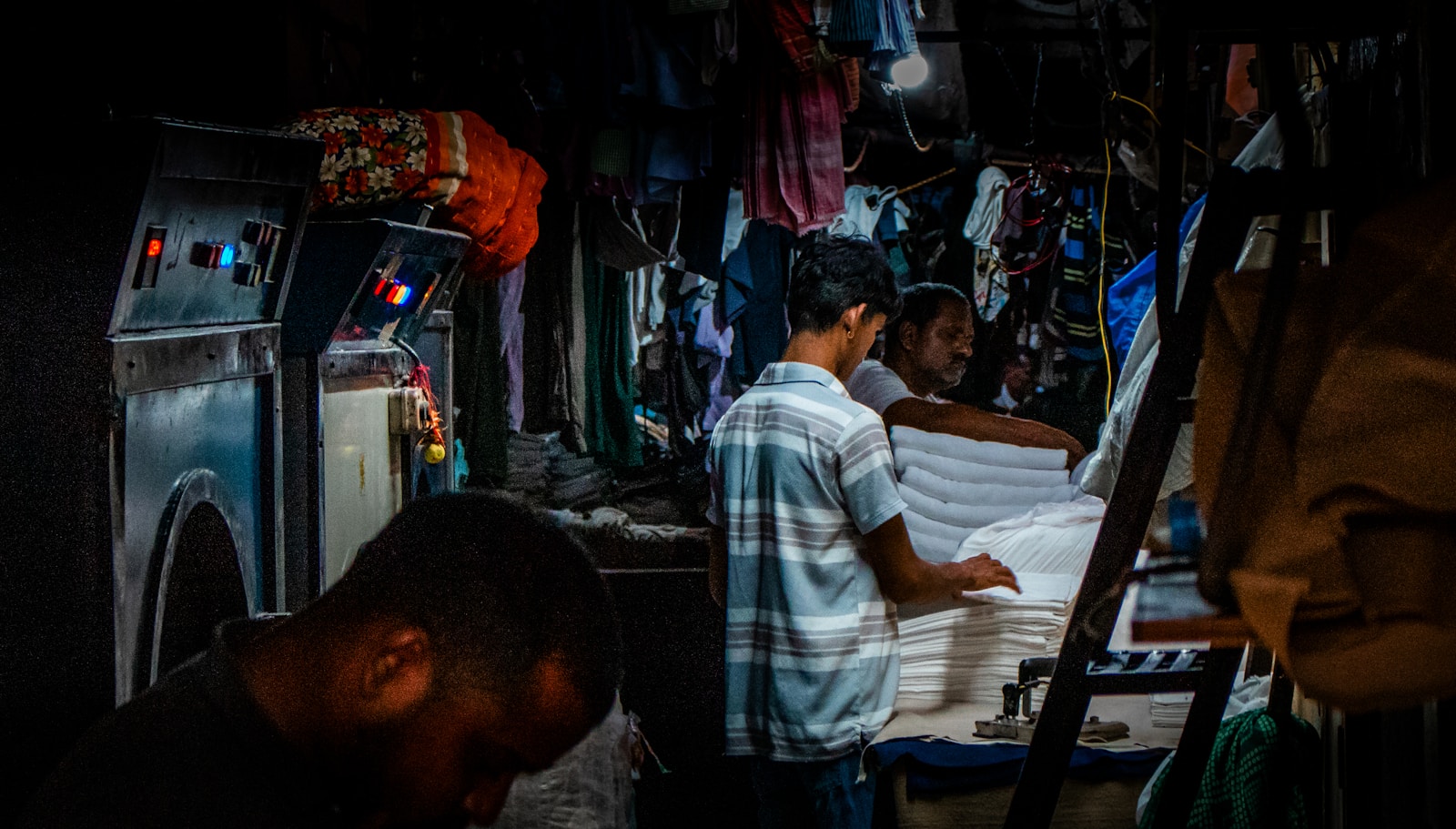 Two workers fold and stack white laundry under a single bare bulb in a dim commercial laundry, pressing machines glowing at the left.