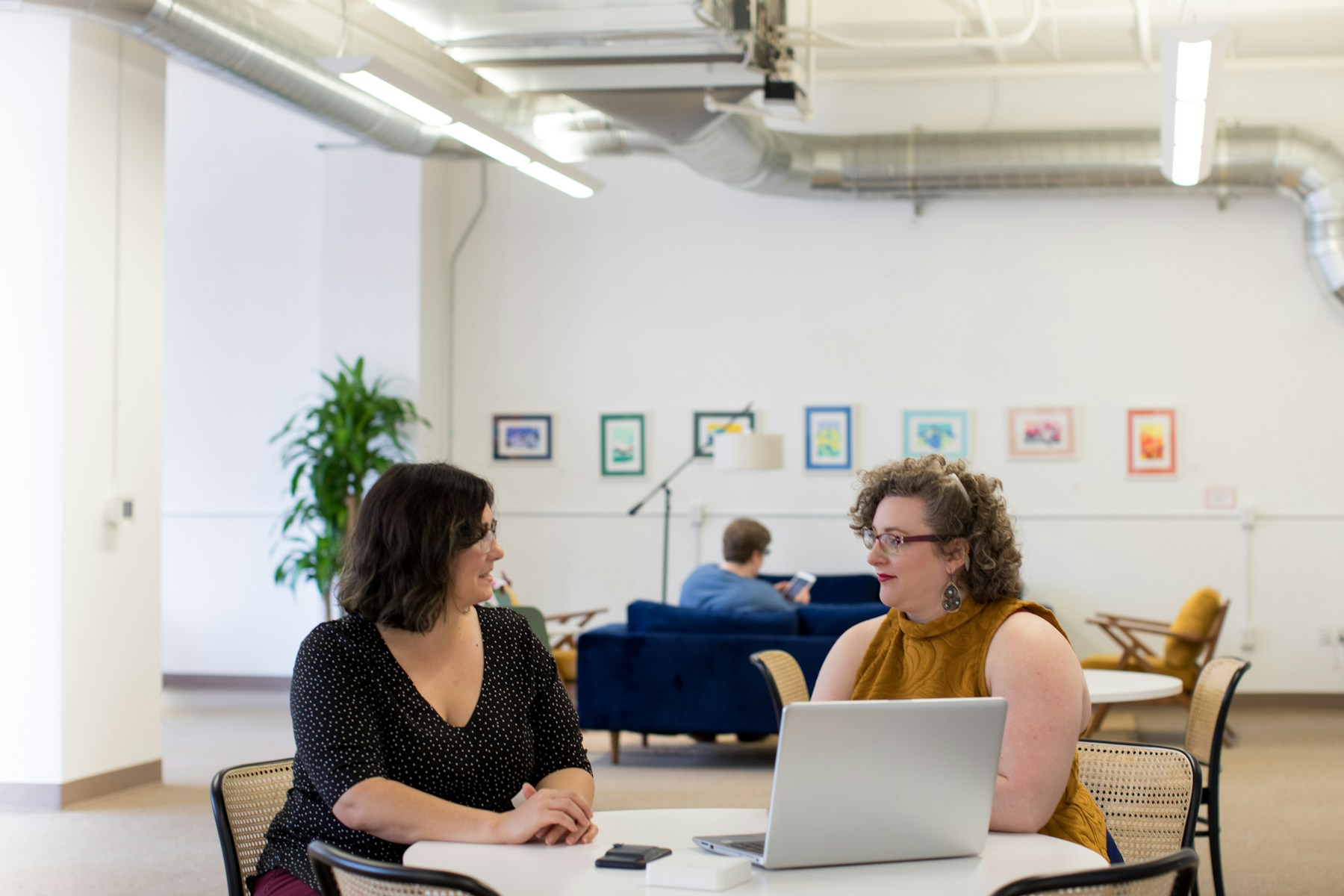 Two women talk across a table with an open laptop in a bright open-plan office with exposed ductwork and framed prints on the wall.